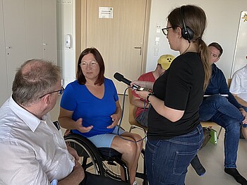 Ein Frau sitzt im Rollstuhl und spricht mit einem Mann während eine andere Frau ein Mikrofon hält.