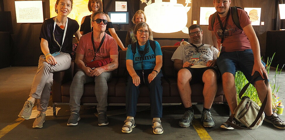 Gruppenfoto mit sieben Personen, die auf einer Couch sitzen und in die Kamera schauen.