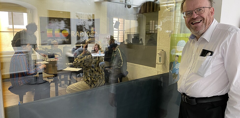 Ein Mann steht vor einem Galsfenster vor dem Radioaufnahmestudio und schaut in die Kamera.