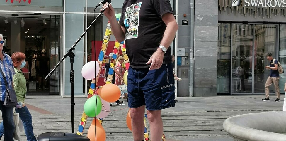 Ein Mann steht auf einer Bühne auf einem öffentlichen Platz und spricht in ein Mikrofon. 
