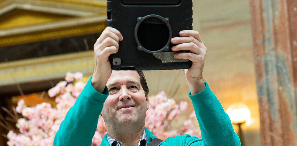 Ein Mann filmt mit einem Tablet die Fotografin und lacht dabei in die Kamera.