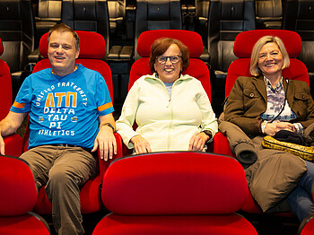 Drei Personen sitzten im Kino auf Kinositzen und lächeln in die Kamera.